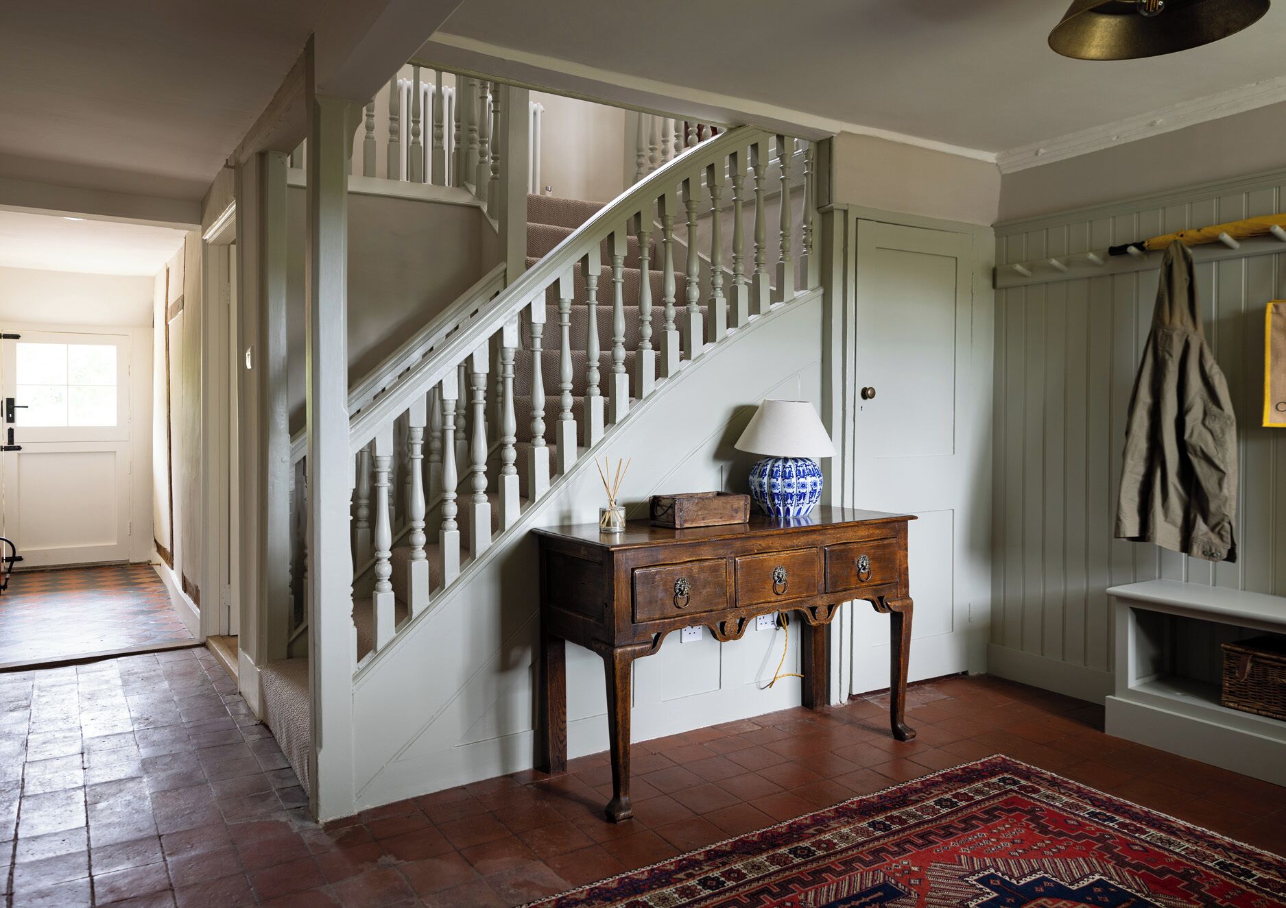 A staircase with white balusters, a wooden console table with drawers, a patterned rug, and a wall-mounted shelf with a jacket.