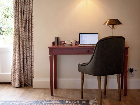 A wooden desk with a laptop, a lamp, and a chair in a room with a patterned curtain and a rug.