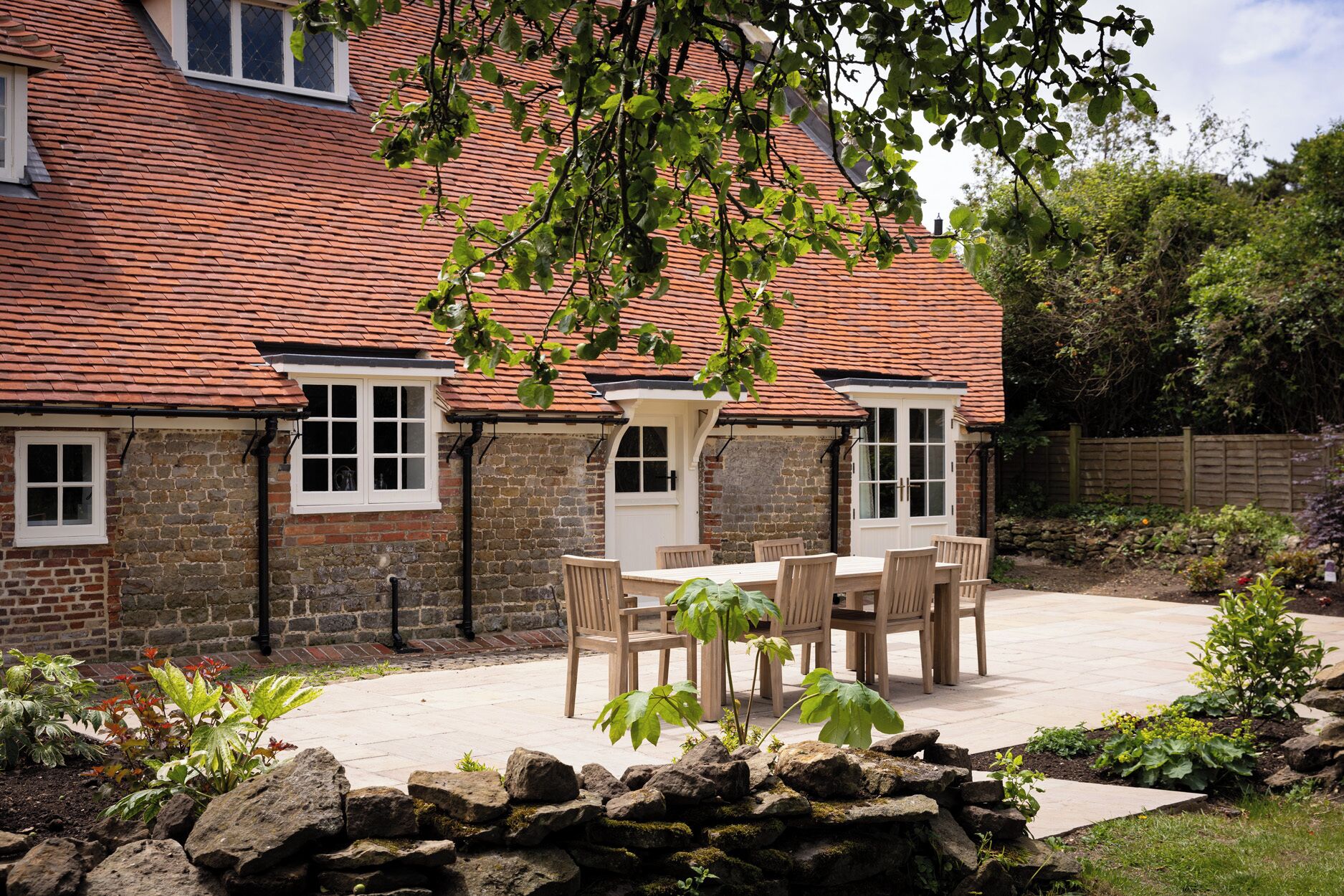 A brick house with a tiled roof, a stone wall, and a garden with a patio table and chairs.