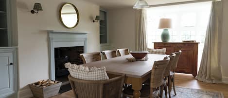 Une salle à manger avec une table en bois, des chaises en osier, une cheminée et un grand miroir au mur.