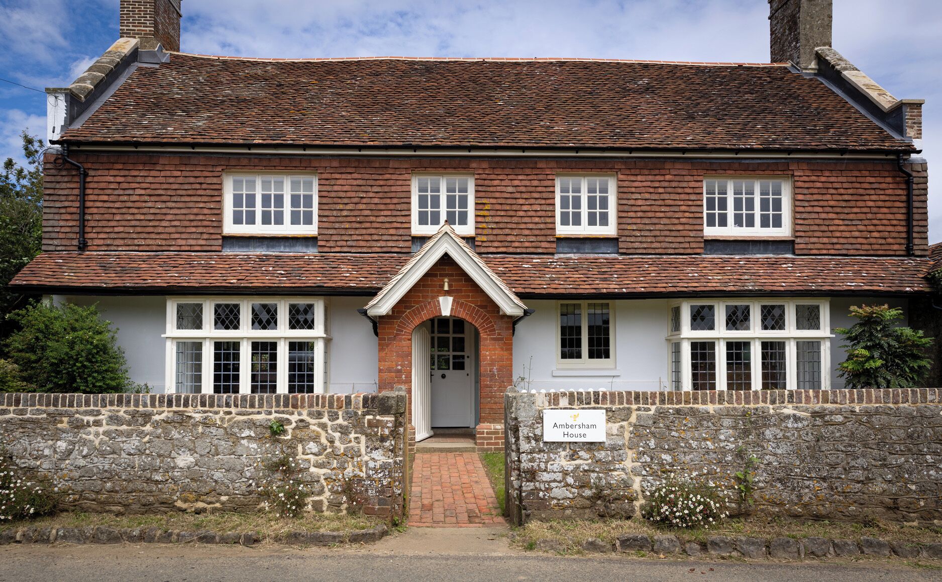 A traditional brick house with a symmetrical facade, a central entrance with a decorative arch, and a stone wall with a sign indicating the property name.