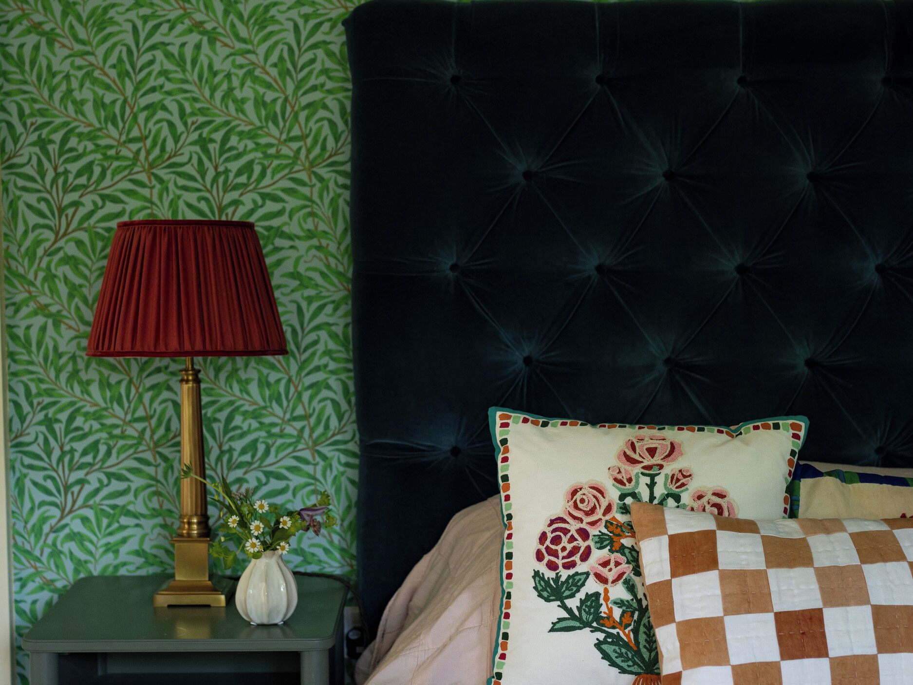A room with a dark upholstered headboard, a bedside table with a lamp and a vase of flowers, and a checkered pillow.