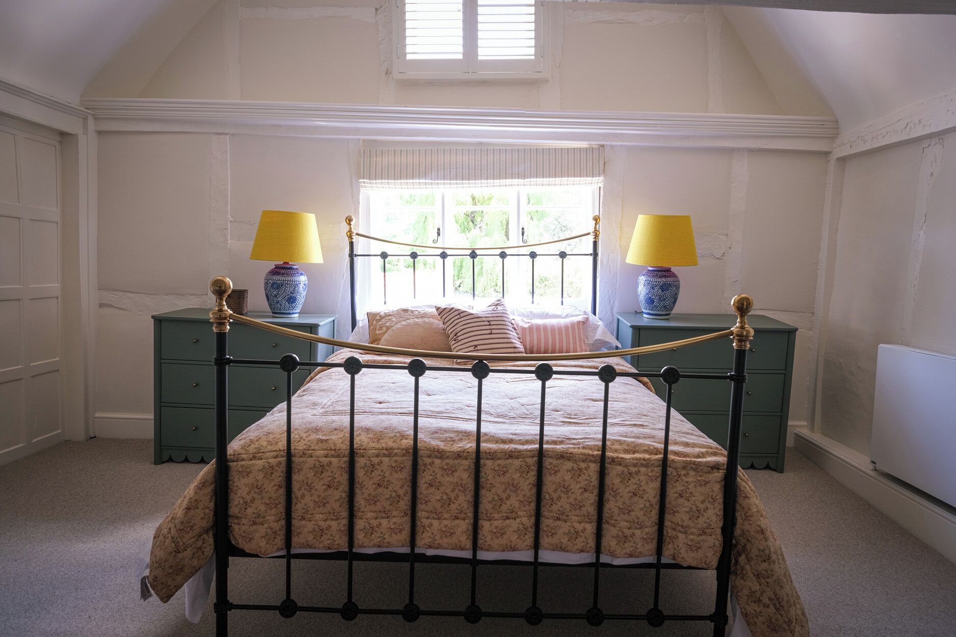 A bedroom with a bed, two bedside tables, a window with blinds, and a radiator.