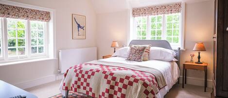 A bedroom with a bed, two bedside tables, a lamp, a radiator, and a window with a view of greenery.