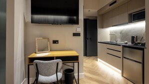 Een moderne hotelkamer met een flatscreen televisie, een houten bureau met stoel, een spiegel, een kleine prullenbak en een kitchenette met een spoelbak en een koelkast.