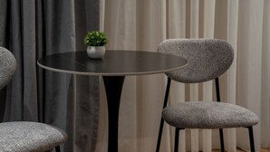 Een ronde tafel met een enkelige plant in pot, twee gestoffeerde stoelen met zwarte poten, en een achtergrond van gordijnen.