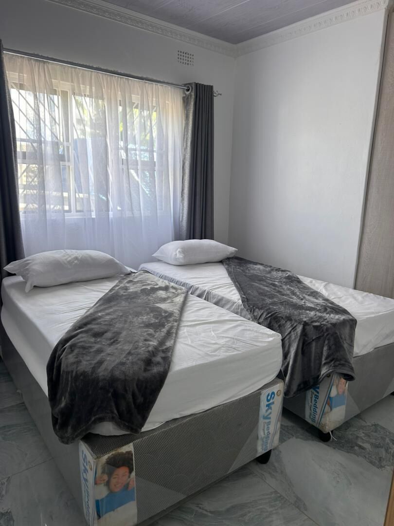 A twin bed with a grey blanket, a window with sheer curtains, and a white wall.