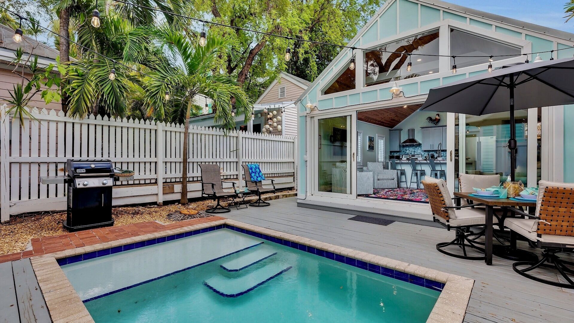 A backyard with a swimming pool, a barbecue grill, outdoor seating, and a house with a large glass door.