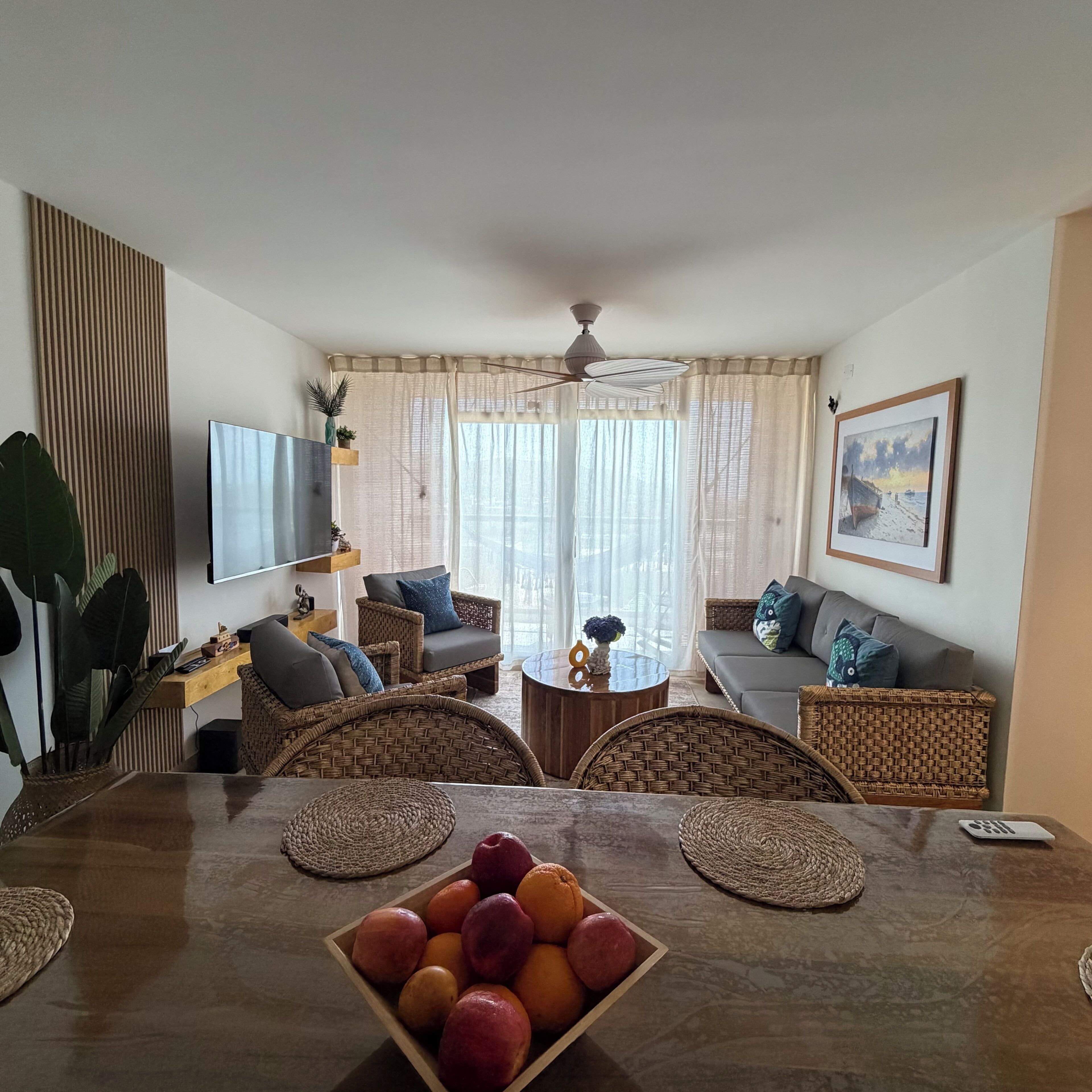 Un salon comprenant une table à manger en bois, des chaises en osier, un canapé, un téléviseur et un tableau accroché au mur.