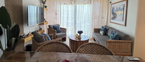 A living room with a wooden dining table, wicker chairs, a sofa, a television, and a painting on the wall.