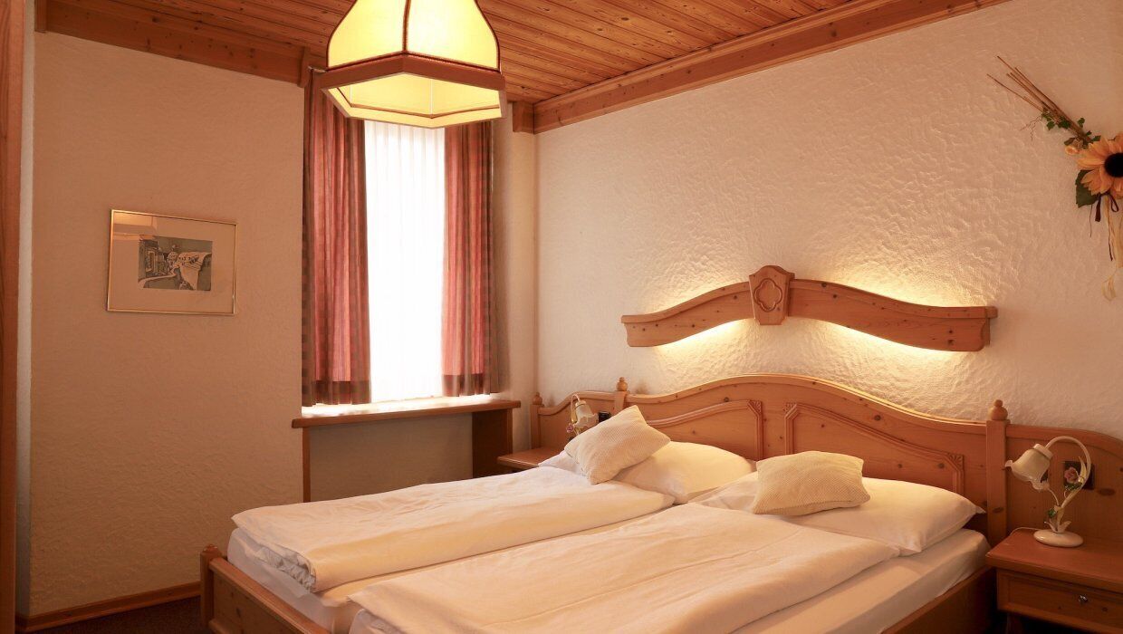 Una cama doble con cabecera de madera, una mesita de noche con lámpara y una ventana con cortinas.