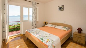 A bedroom with a bed, a nightstand, a lamp, a picture on the wall, and a view of the sea through a window. - Apartments Stipo - Comfort Two Bedroom Apartment With Balcony and Sea View (Janjina)