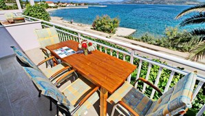 A wooden table with chairs on a balcony overlooking a coastal area with a clear view of the sea and distant mountains. - Apartments Stipo - Comfort Two Bedroom Apartment With Balcony and Sea View (Janjina)