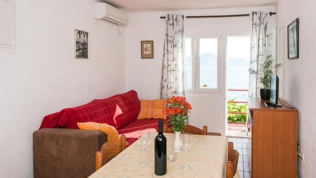 A room with a dining table, a red sofa, a TV on a wooden cabinet, and a view of the sea through a window. - Apartments Stipo - Comfort Two Bedroom Apartment With Balcony and Sea View (Janjina)