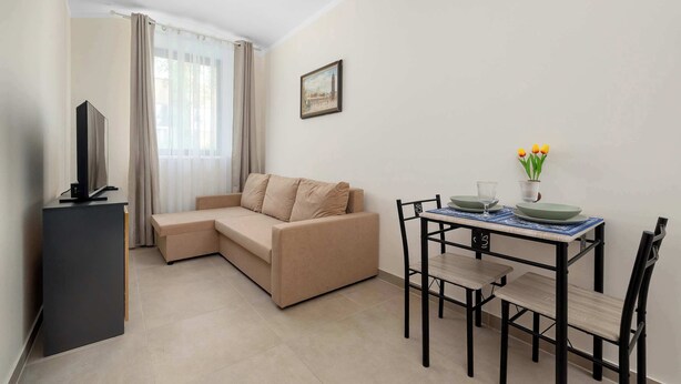 Una sala de estar con un sofá, una mesa de comedor con sillas, un televisor sobre un mueble bajo y una ventana con cortinas.
