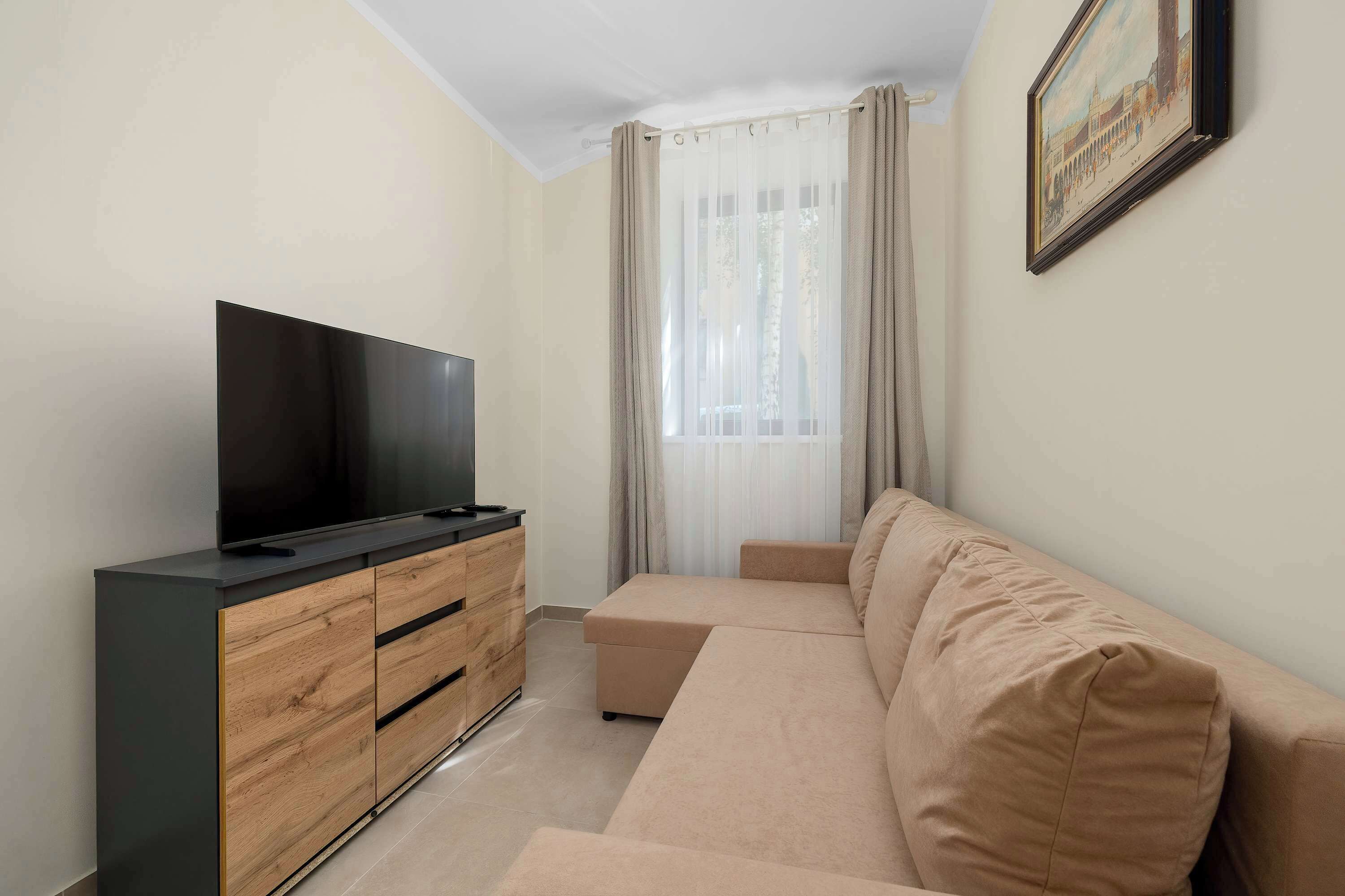 Een moderne woonkamer met een flatscreen televisie op een houten kast, een beige bank en een groot raam met transparante gordijnen.