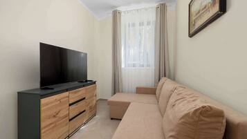 Een moderne woonkamer met een flatscreen televisie op een houten kast, een beige bank en een groot raam met transparante gordijnen.