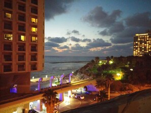 Un paisaje urbano costero al atardecer con edificios iluminados, un puente y una playa.