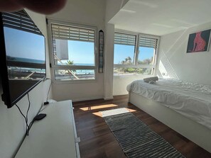 A bedroom with a bed, a desk, a TV, and large windows with a view of the ocean. - Red Gordon Beach View Apartments + Bomb Shelter (Tel Aviv-Yafo)
