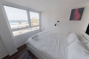 A bedroom with a large window, a bed with white linens, a wall-mounted lamp, and a piece of abstract art. - Red Gordon Beach View Apartments + Bomb Shelter (Tel Aviv-Yafo)