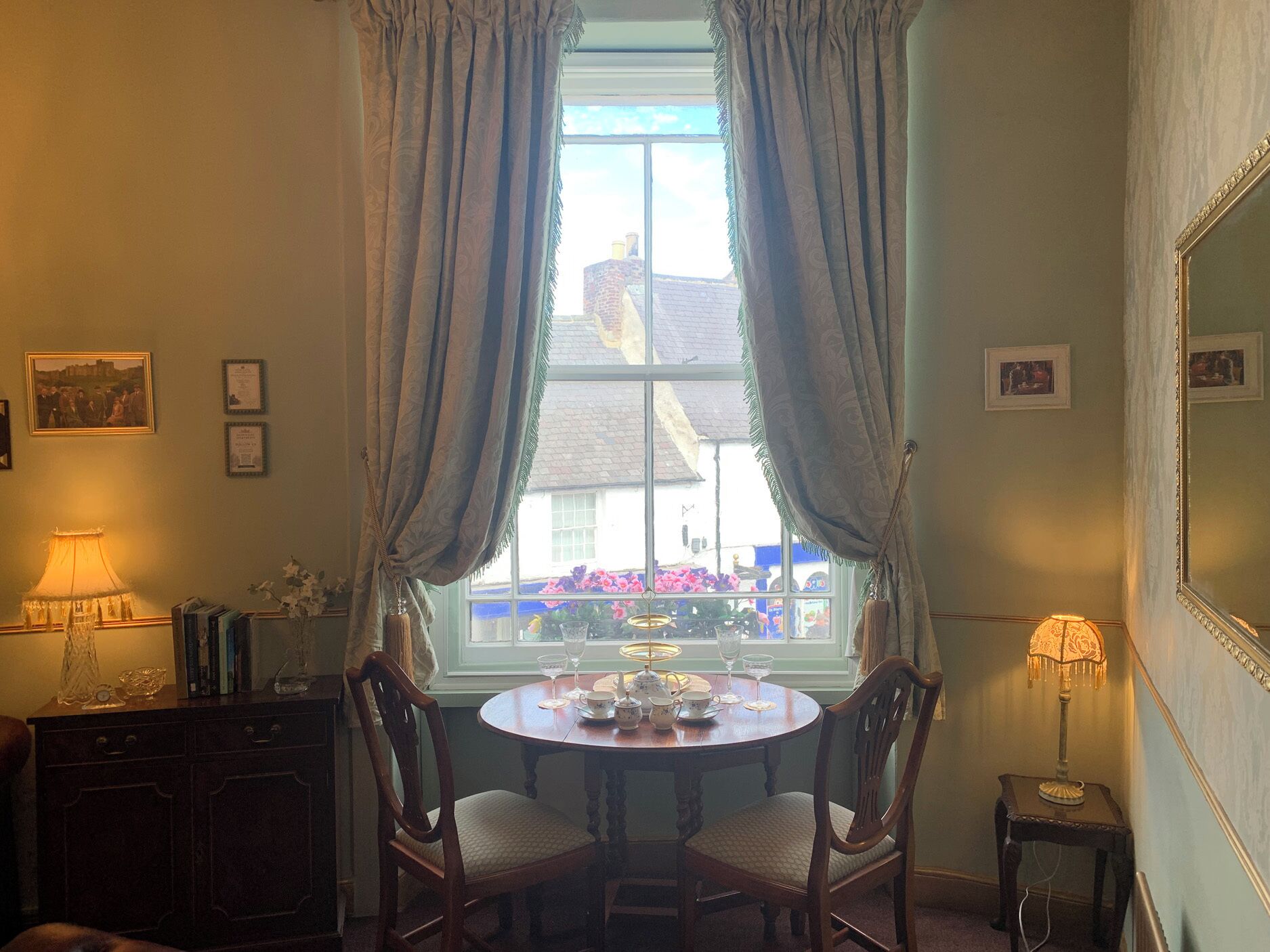 A room with a round table set for tea, two chairs, a window with curtains, a lamp, and framed pictures on the wall.