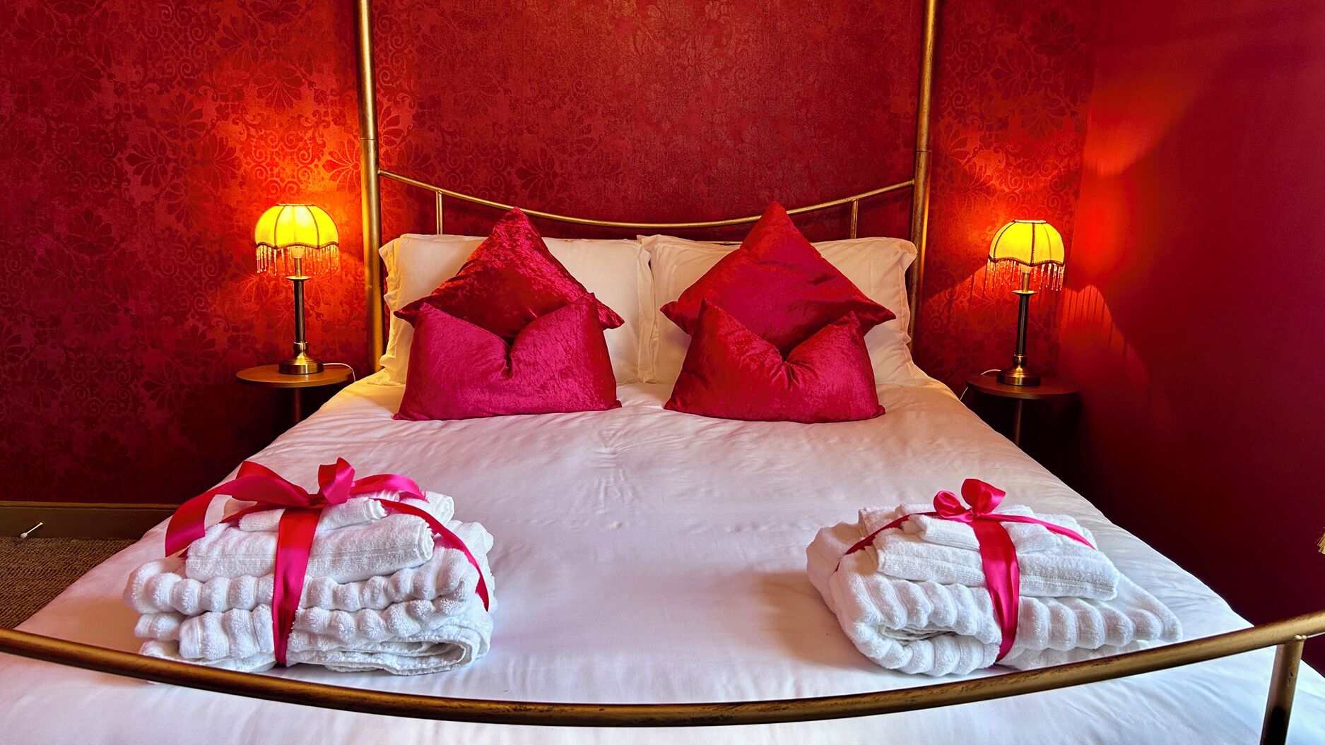 A hotel room with a bed, two red pillows, two bedside lamps, and two towels with pink ribbons.