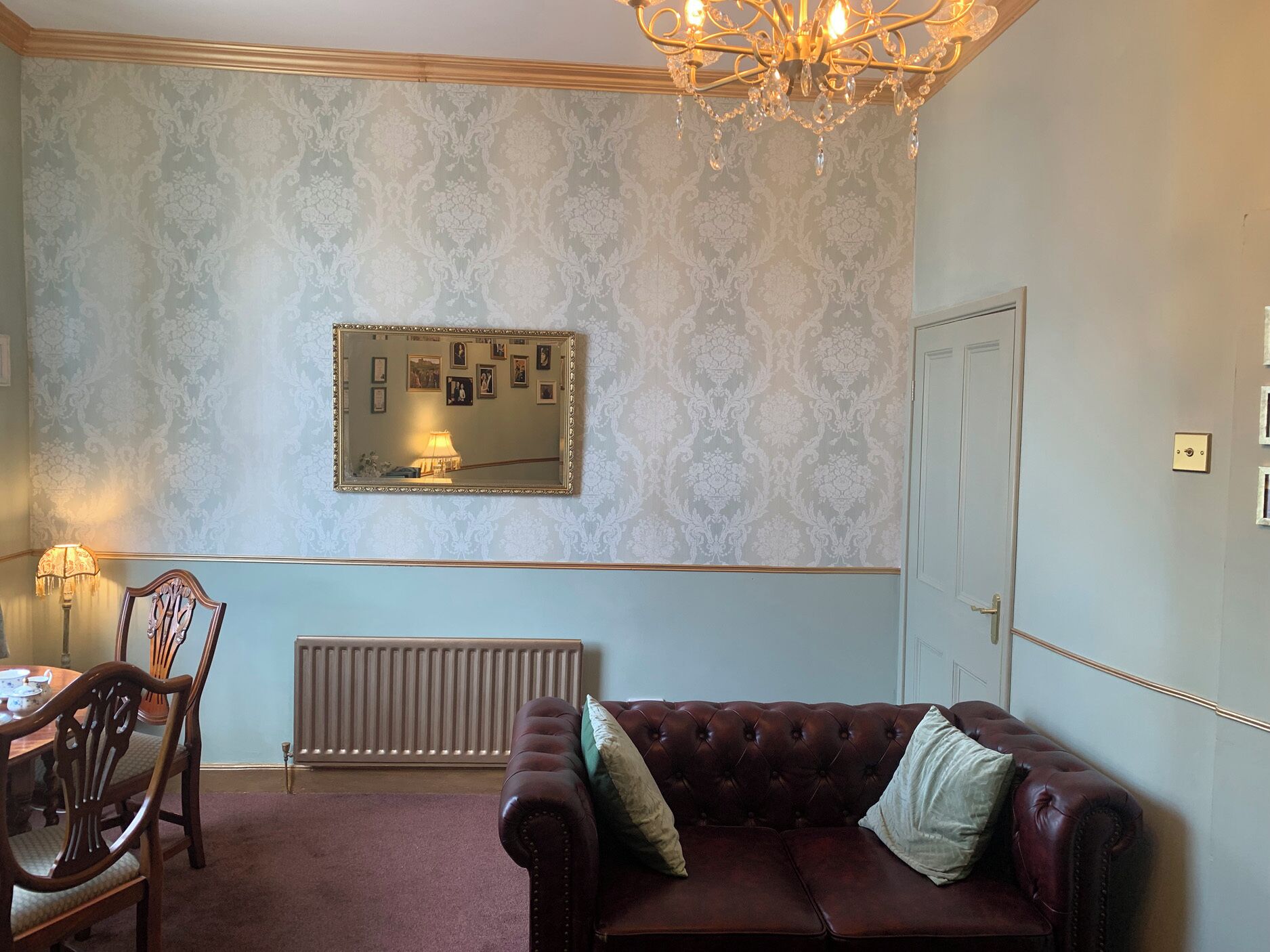 Une pièce avec un radiateur, un canapé en cuir marron agrémenté de coussins, une table à manger en bois avec des chaises, un miroir mural orné de tableaux et un lustre.