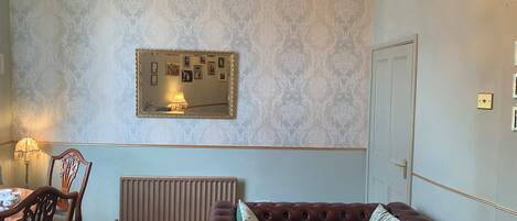 Une pièce avec un radiateur, un canapé en cuir marron agrémenté de coussins, une table à manger en bois avec des chaises, un miroir mural orné de tableaux et un lustre.
