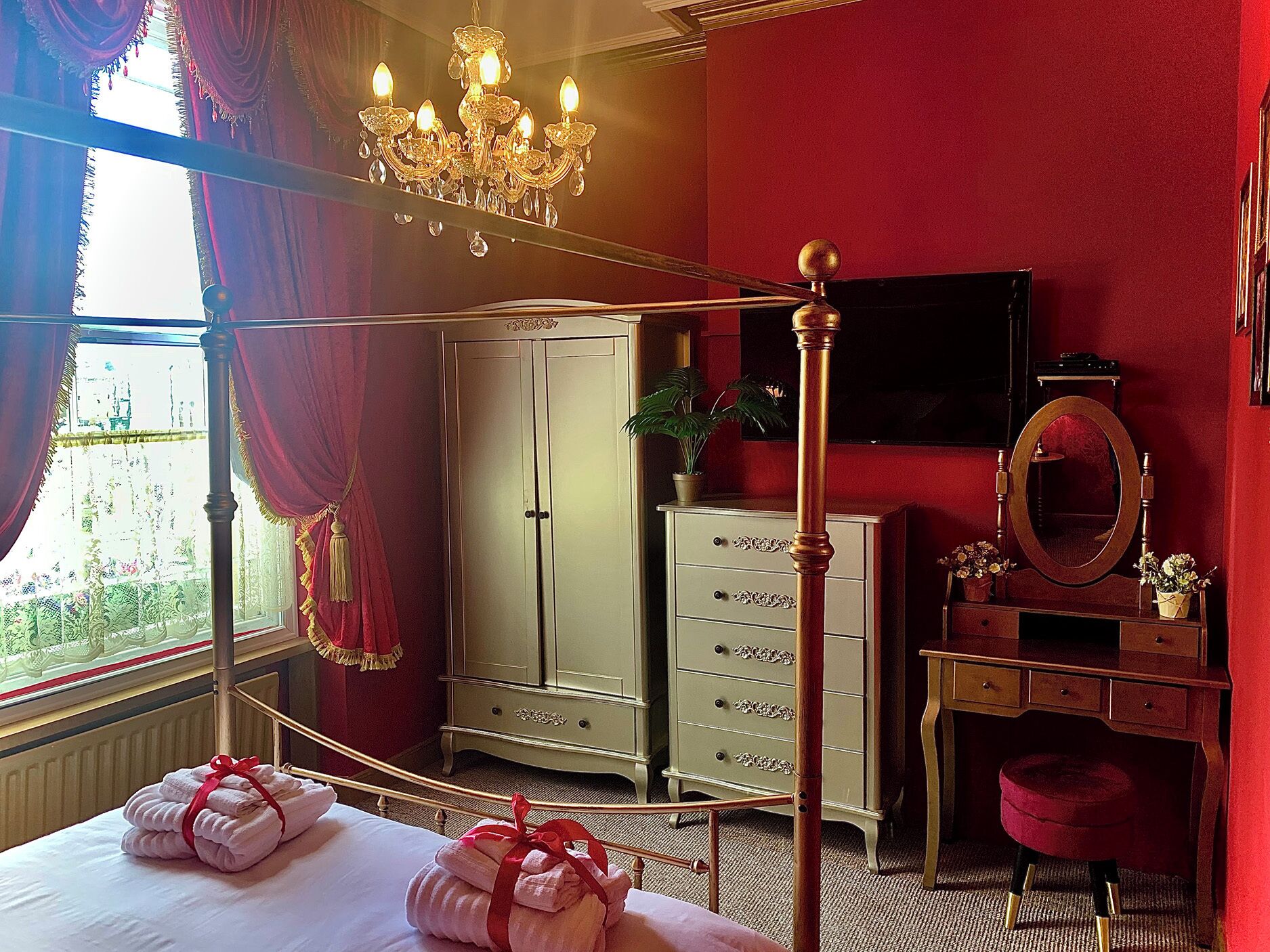 A hotel room with a four-poster bed, a wardrobe, a vanity with a mirror, and a television on the wall.