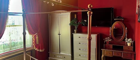 Een hotelkamer met een hemelbed, een kledingkast, een kaptafel met spiegel en een televisie aan de muur.