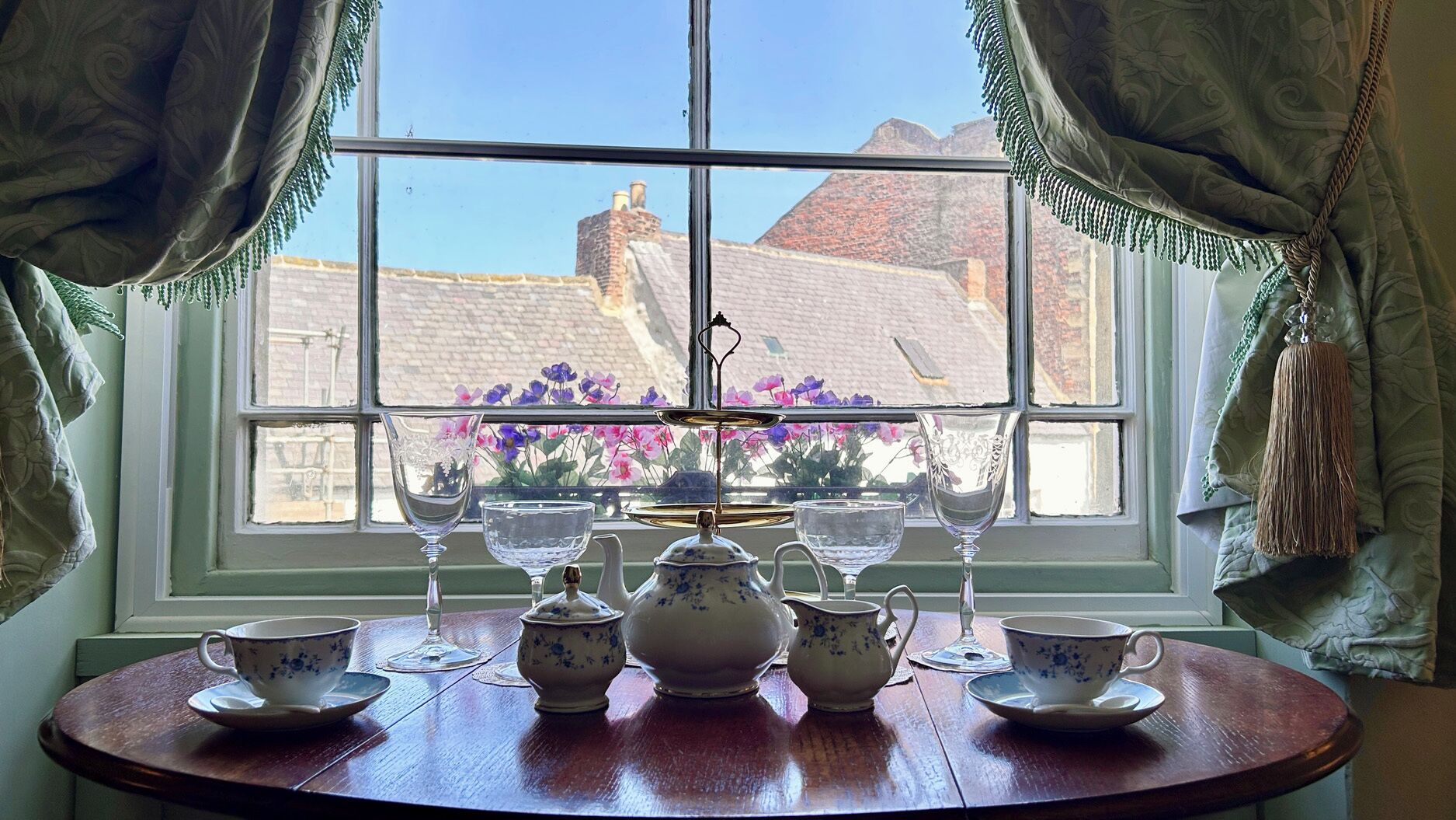 A table set with tea service and glassware in front of a window with curtains.