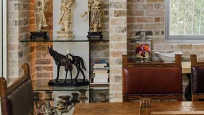 Une pièce avec un mur en briques, une étagère en verre où sont exposées des statues, une table en bois et une fenêtre donnant sur un paysage.