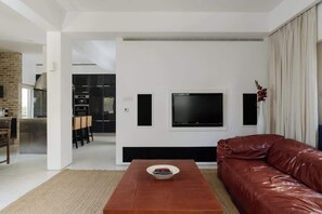 A modern living room with a leather sofa, a coffee table, a flat-screen TV mounted on the wall, and a dining area visible in the background. - Gold Gordon Beach Apartments - Chefs apartment (Tel Aviv-Yafo)