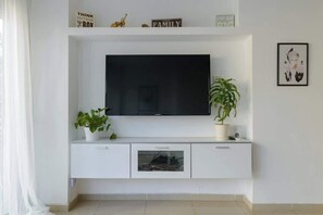 A white entertainment unit with a flat-screen TV, two potted plants, and a framed picture on the wall. - Green Gordon Beach View Apartments + Bomb Shelter (Tel Aviv-Yafo)