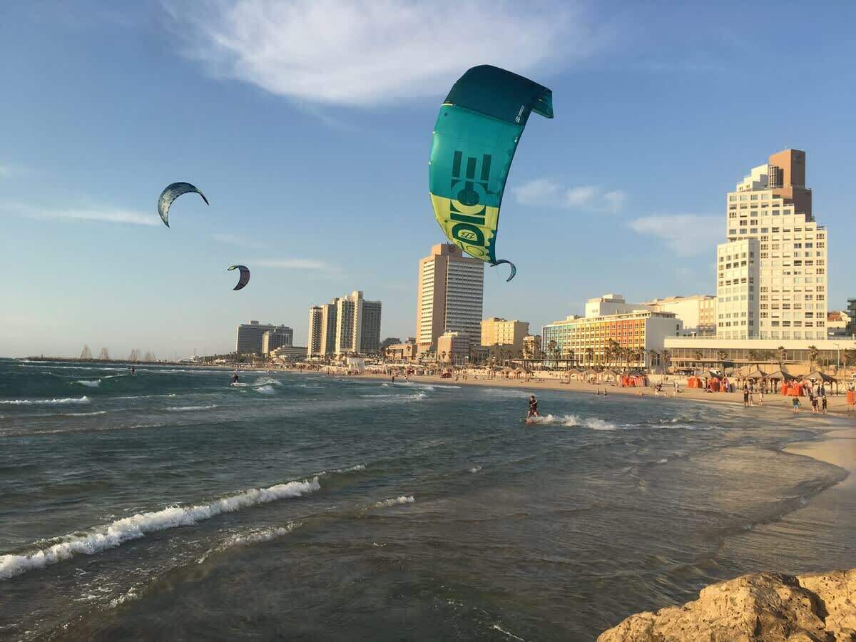 Una playa con olas, el horizonte de una ciudad y cometas en el cielo.