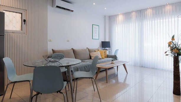 Een moderne woonkamer met een bank, een glazen salontafel en stoelen. Er is een raam met gordijnen, een airconditioning en een ingelijst schilderij aan de muur.