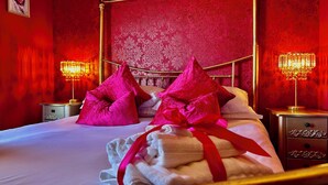 A bed with pink pillows, a red bow, and a white towel, set against a red patterned wall with two bedside lamps. - Downton II (Alnwick)