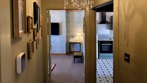A hallway with a chandelier, framed pictures, a small table with a lamp, and a patterned floor leading to a room with a stove. - Downton II (Alnwick)