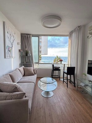Ein modernes Wohnzimmer mit einer Couch, einem Glastisch, einem Essbereich mit Stühlen und Blick auf das Meer.