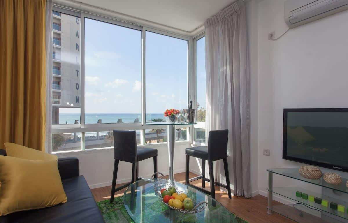 Un salon moderne avec un canapé, deux fauteuils, une table basse en verre sur laquelle sont disposés des fruits, un téléviseur et une vue sur la mer.