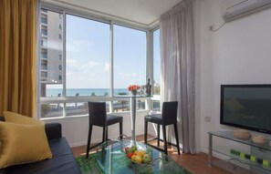 Ein modernes Wohnzimmer mit einer Couch, zwei Sesseln, einem Glastisch mit Obst, einem Fernseher und Meerblick.