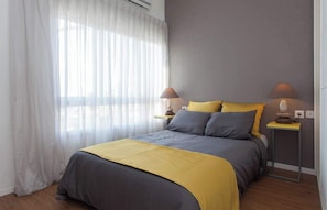 Ein Schlafzimmer mit einem Bett, zwei Nachttischlampen, einem Fenster mit durchscheinenden Vorhängen und einem Nachttisch.
