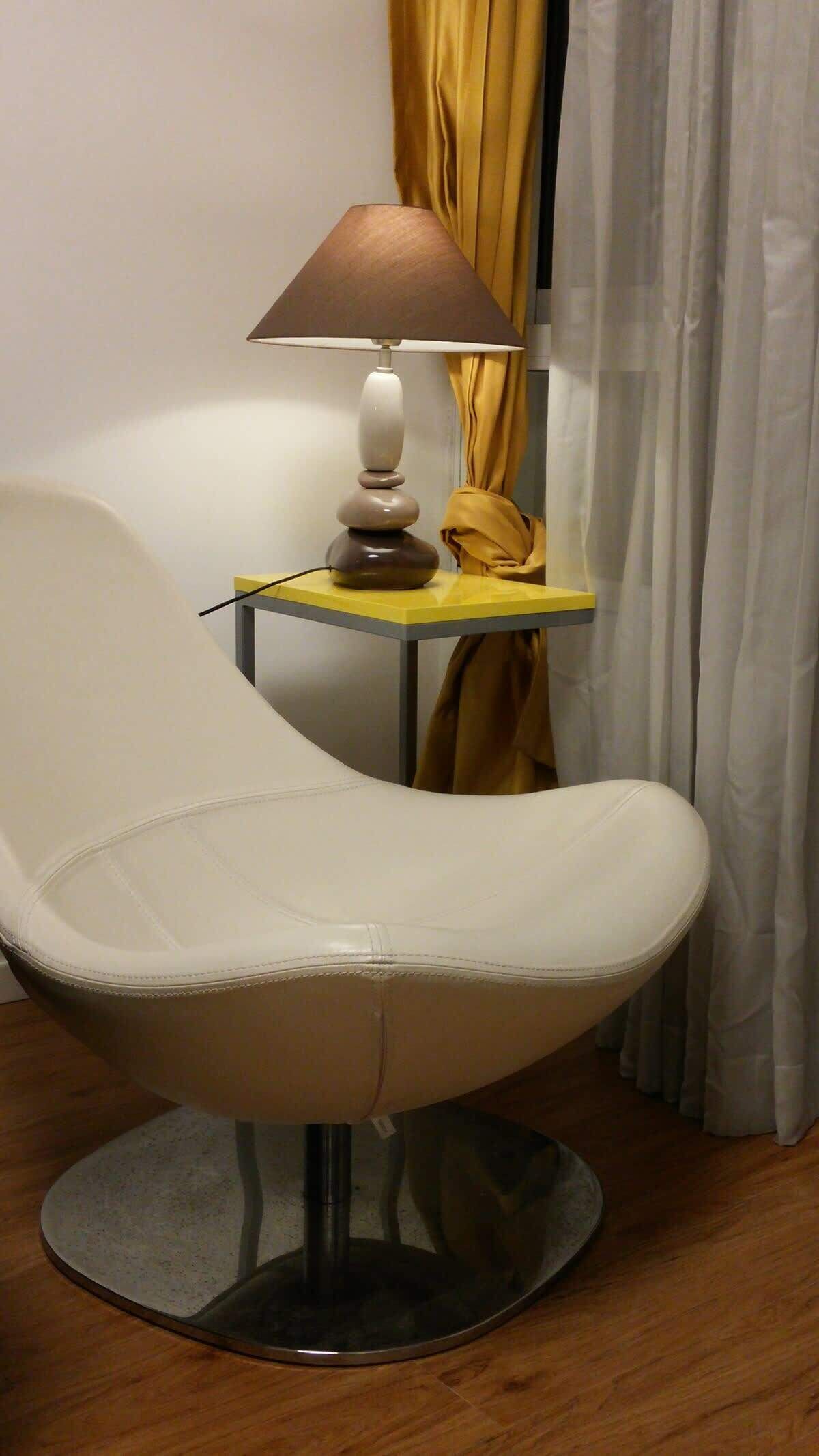 A modern chair with a round base, a small table with a lamp, and a curtain in the background.