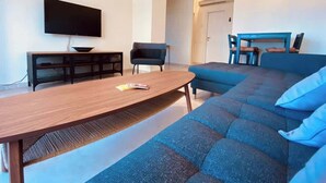 Ein modernes Wohnzimmer mit Couchtisch aus Holz, einem blauen Sofa, einem schwarzen Sessel und einem flachbildfernseher an der Wand.