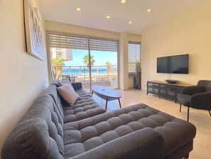 A living room with a large sectional sofa, a flat-screen TV, and a balcony with a view of the sea. - White Gordon Beach Apartment + Bomb Shelter (Tel Aviv-Yafo)