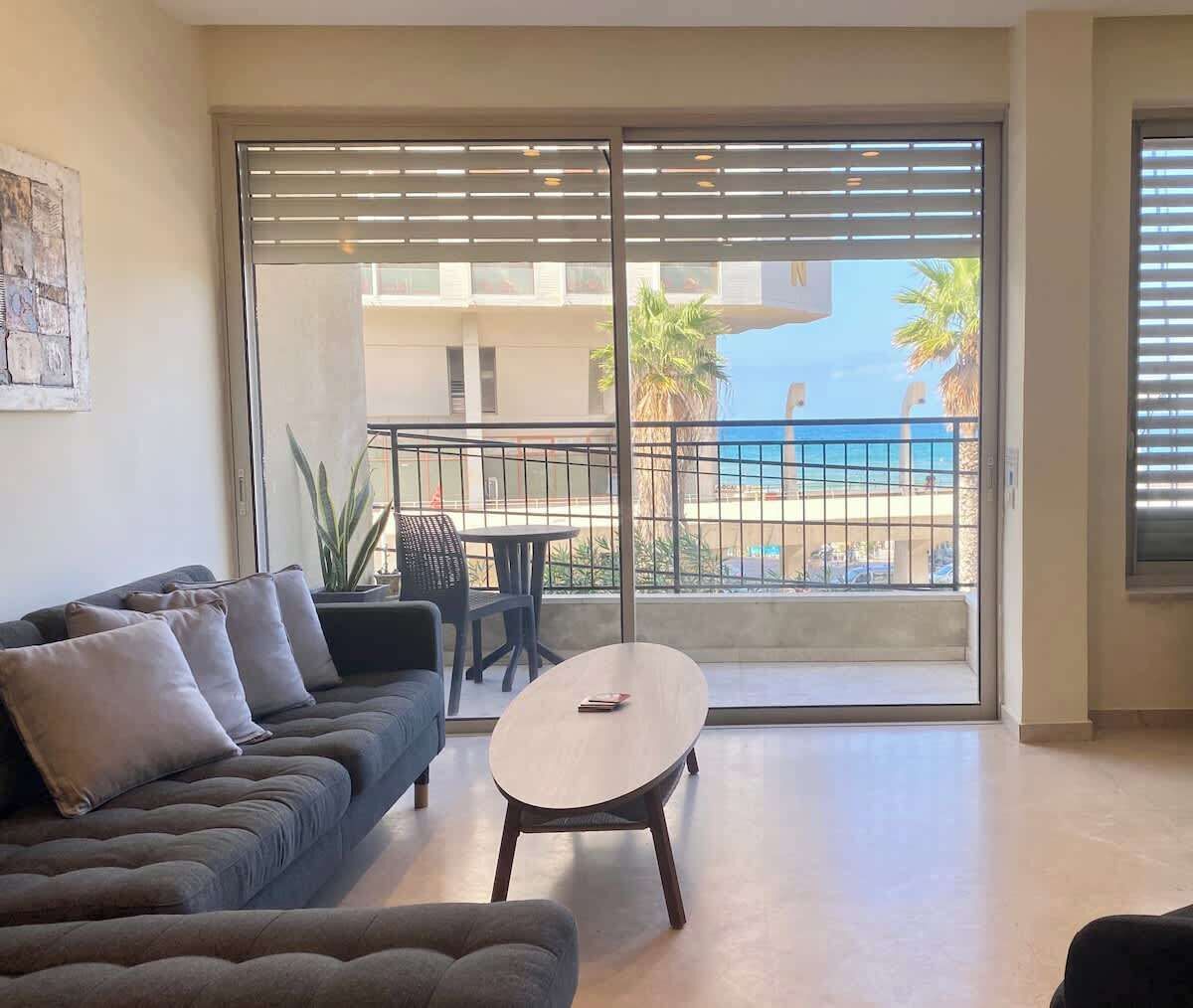 Een woonkamer met een bank, een bijzettafeltje en uitzicht op een balkon met een hekwerk en palmbomen.