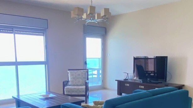 A living room with a blue sofa, a wooden coffee table, a TV stand, and a chair. There is a large window providing natural light. - Krystal View Family 2 parking Bomb Shelter (Bat Yam)