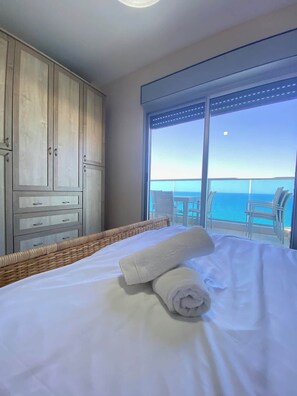 Quarto de hotel com uma cama, uma mesinha de vime e vista para o oceano através de uma janela grande.