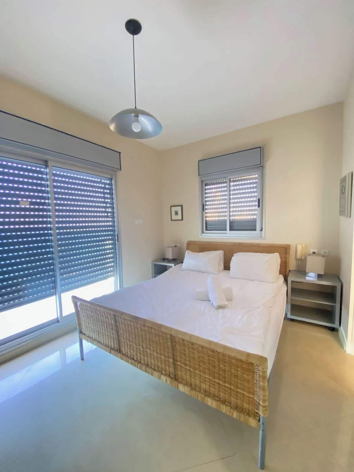 A bedroom with a bed, a nightstand, a lamp, a ceiling light, and a window with blinds.