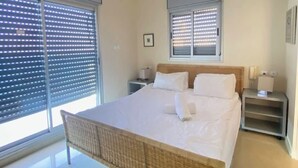 A bedroom with a bed, a nightstand, a lamp, a ceiling light, and a window with blinds. - Krystal View Family 2 parking Bomb Shelter (Bat Yam)